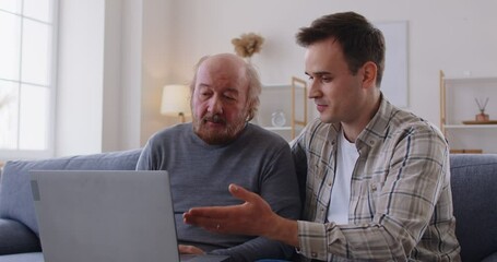 Senior father and an adult son watching a video together. Old man together with his young son sitting on the couch, looking at a laptop, watching and discussing a movie. Family, leisure concept - Powered by Adobe