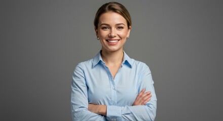 Confident young blonde woman with blue eyes smiling at the camera, standing with arms crossed against a gray background. She is wearing a light blue professional shirt.