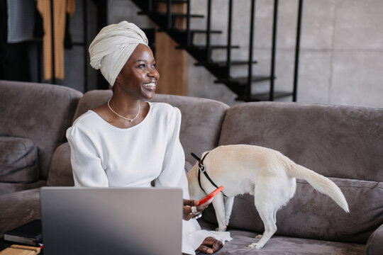 A smiling woman in white enjoys the company of her dog while engaging with her laptop in a bright, stylish living space.