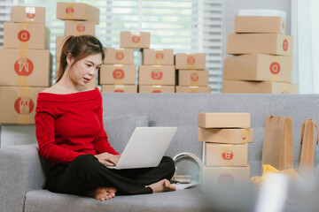 A young woman is comfortably working on a laptop on a sofa, surrounded by numerous shipping boxes,...
