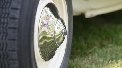 Polished chrome hubcap on a vintage car whitewall wheel reflecting green lawn at an outdoor auto show.