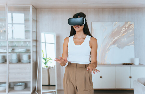 A woman in a white tank top and beige pants enjoys a virtual reality experience, smiling and immersed in a bright, modern living room.