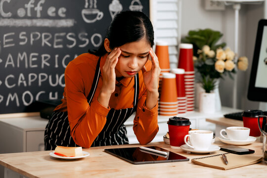 A young Asian female barista in a striped apron and orange shirt stands in a cozy modern café, looking sad, stressed, and disappointed, feeling exhausted and overwhelmed by business loss and failure.
