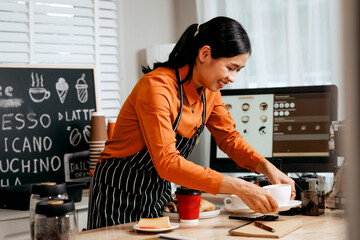 A young Asian female barista in a striped apron and orange shirt stands in a cozy modern café,...