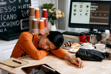 A young Asian female barista in a striped apron and orange shirt stands in a cozy modern café,...