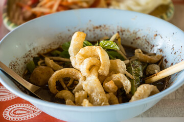 Close-up of a traditional Asian noodle soup with crispy pork rinds and fresh herbs served in a light blue bowl on a patterned cloth.
