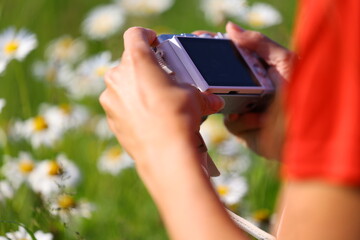Female hands with camera capturing close-up of wild daisies in meadow, wearing red shirt among blooming summer flowers