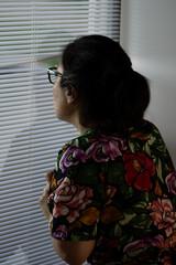A woman observes what is happening outside, hiding behind the blinds. The scene creates a sense of mystery and intrigue.