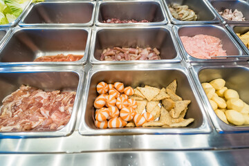 Metal trays filled with various fresh food ingredients in a buffet display, including meat, seafood, and assorted processed items