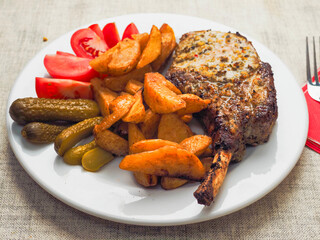 A plate of food with a premium pork chop in the bone, wedges potatoes, and tomatoes. The plate is set on a table with a fork and a knife. Fine tasty dinner with high quality meat.