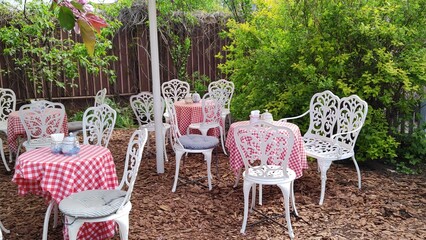 Cozy outdoor cafe with delicate white chairs and tables with red checkered tablecloths on lawn or in garden. Metal furniture in classic style. Openwork white metal chair in cafe