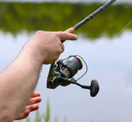 Spinning fishing on the river, fisherman holds a fishing rod in his hands and turns the reel, blurred background. Male hands hold a spinning rod against the background of a river