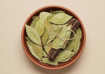 Overhead view of bay leaves and a bundle of sticks in a brown ceramic bowl on a beige background