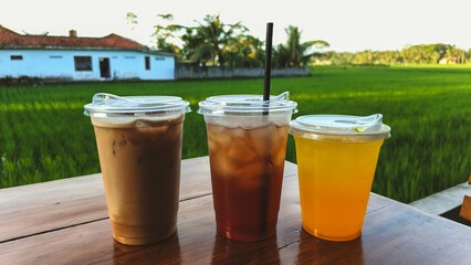 Three refreshing iced drinks—coffee, tea, and citrus—served on a wooden table with a peaceful green rice field in the background. A perfect countryside café moment.