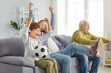 Happy multi generation excited male family sitting on sofa, cheering their favorite football team. Father, son and grandfather watching soccer on TV at home, raising hands up, celebrating scored goal.