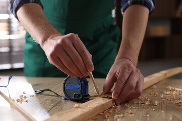 Man measuring plank with tape and pencil at wooden table indoors, closeup