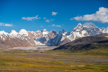 A picturesque scene of towering snow-capped mountain peaks and glaciers, overlooking a wide, green...