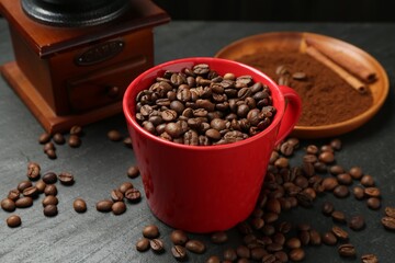 Aromatic coffee beans in cup and grinder on black table, closeup