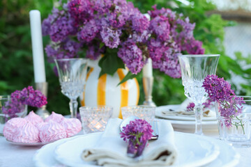 Beautiful lilac flowers, zephyrs, plates, cutlery, glasses and burning candles on table in garden, closeup
