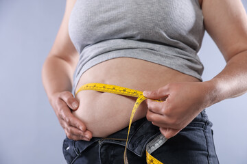 Overweight woman measuring her belly with tape on light background, closeup