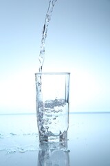 Pouring water into glass at mirror table against light blue gradient background, closeup