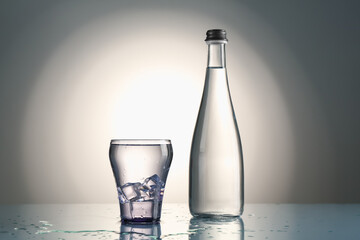 Fresh water with ice cubes in glass and bottle on mirror table against gray gradient background