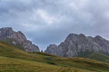 A dirt track winds through verdant green hills, leading towards a dramatic range of rugged, rocky...