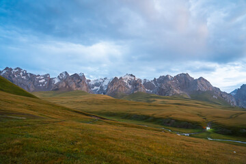 A wide landscape view of a vast mountain valley at twilight, with golden and green fields, a small...