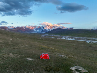An eye-level view of a vibrant red camping tent pitched in a vast green field, with majestic...