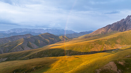 Sunlight illuminates rolling golden and green mountain slopes, with a faint rainbow visible in the...
