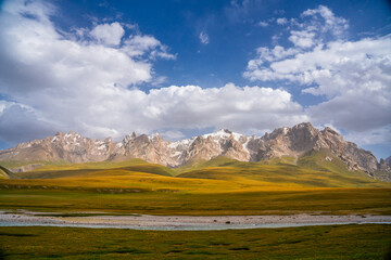 A wide landscape of a river flowing through golden and green fields, with a distant range of...