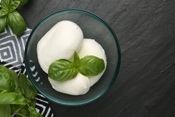 Pieces of delicious mozzarella cheese and basil in bowl on dark textured table, flat lay. Space for...