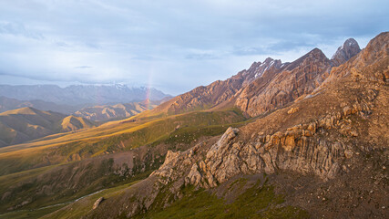 A dramatic landscape featuring a rugged mountain range, some slopes bathed in golden light, and a subtle rainbow in the distant hazy valley