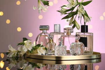 Many different perfume bottles and flowers on mirror surface against pink background with blurred...