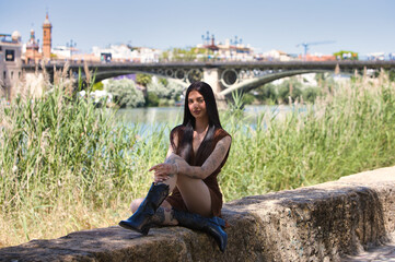 Latin woman, young and beautiful brunette with tattoos on her arms and legs, wearing a brown dress and black boots, sitting on a stone wall. In the background is the famous bridge in Seville, Spain.