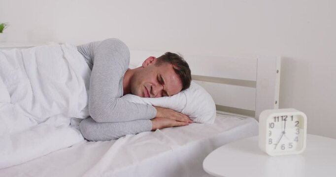 Sleepy young man sleeping on bed in bedroom at home with alarm clock on table and cant waking up. Male person trying to wake up in the morning indoors. 4k video. Slow motion video.