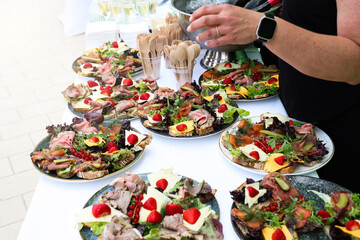 Catering table with assorted open-faced sandwiches, cheese, meat, fruits, and wooden cutlery, prepared for a festive outdoor event or celebration.