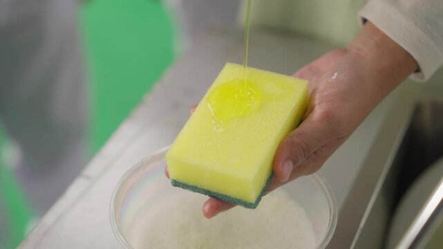 Close-up shot of a woman's hand pouring dish detergent onto yellow sponge. Closeup of woman using dishwashing detergent for wash up dishes.