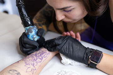 Close-up of a tattoo artist creating a floral tattoo on a woman's hand using a tattoo machine in...