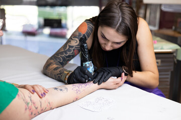 Close-up of a tattoo artist creating a floral tattoo on a woman's hand using a tattoo machine in...