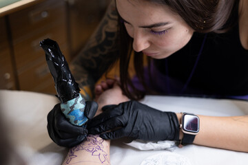 Close-up of a tattoo artist creating a floral tattoo on a woman's hand using a tattoo machine in...