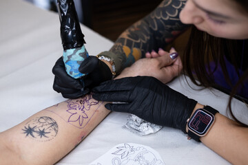 Close-up of a tattoo artist creating a floral tattoo on a woman's hand using a tattoo machine in...
