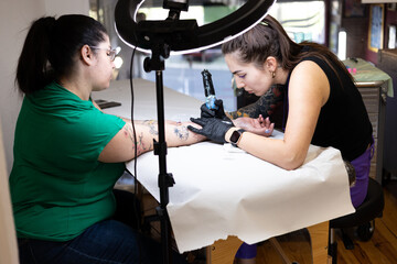 Close-up of a tattoo artist creating a floral tattoo on a woman's hand using a tattoo machine in...