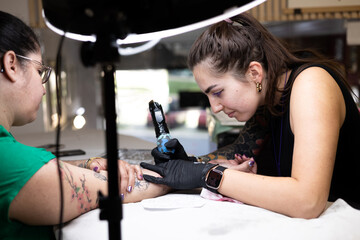 Close-up of a tattoo artist creating a floral tattoo on a woman's hand using a tattoo machine in...