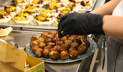 Chef wearing black gloves prepares meatballs on skewers for a catering event in a professional kitchen. Tasty food served on a ceramic plate, ready for guests.