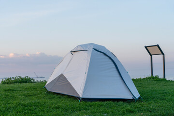 Camping tent set up on green grass near the water during a tranquil evening with soft skies and distant views of ships on the horizon