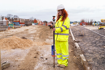 Portrait of confident female land surveyor with long hair in bright safety gear using surveying...