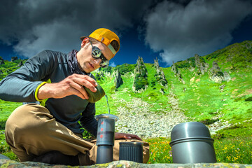 Male backpacker preparing coffee with Aeropress next to camping stove while sitting on rock in...
