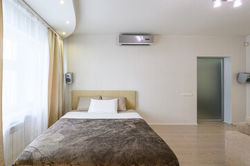 cozy bedroom with a bed, beige headboard, warm-toned bedding, sheer curtains, and an air conditioner on a white wall. A door with frosted glass is visible