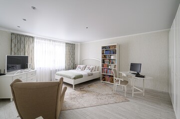 bright bedroom features a bed, desk, TV, and beige armchair. The room has a white and beige color...